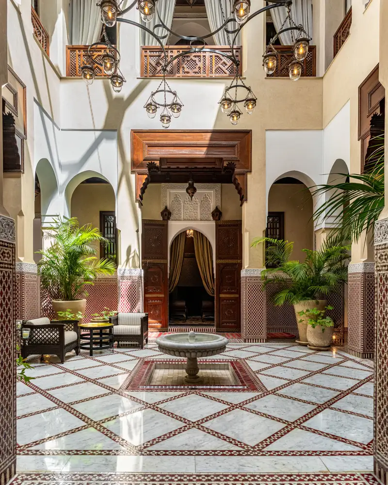 Patio de riad avec fontaine centrale, sol en zellige, plantes et porte en bois sculpté