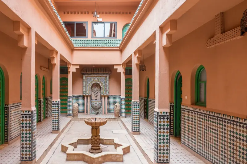 Galerie aux murs roses autour d’une fontaine en zellige